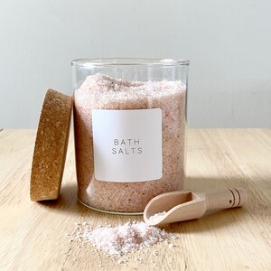 May include: A glass jar filled with pink bath salts with a white label that reads "Bath Salts". A wooden scoop and a cork lid are next to the jar.