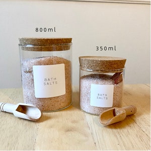 May include: Two glass jars with cork lids filled with pink bath salts. The jars have white labels that say "Bath Salts". The larger jar is 800 ml and the smaller jar is 350 ml. There are two wooden scoops next to the jars.