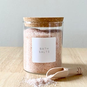 May include: A glass jar with a cork lid filled with pink bath salts. The jar has a white label that says "Bath Salts". A wooden scoop is next to the jar.