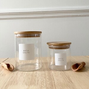 May include: Two glass jars with bamboo lids and labels that say "Bath Salts". The jars are on a wooden surface with two wooden scoops.