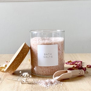 May include: A glass jar filled with pink bath salts with a white label that reads "Bath Salts". The jar has a cork lid and is sitting on a wooden surface with a wooden scoop next to it.