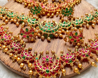 May include: A traditional Indian jewelry set featuring a gold necklace with multiple crescent moon pendants adorned with pink and green gemstones. The set also includes matching earrings.