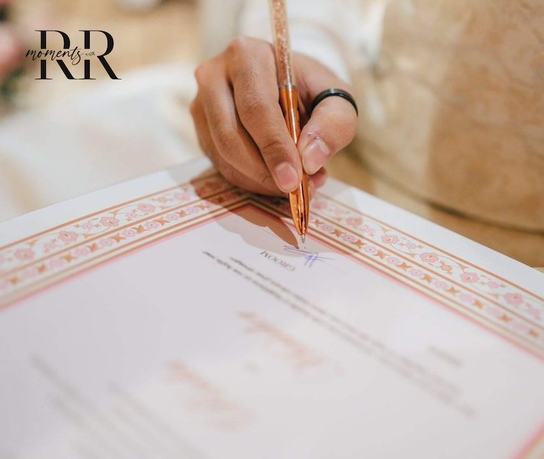 May include: A person signing a document with a rose gold pen. The document has a pink and gold border with a floral pattern. The text "moments..." is visible in the top left corner of the image.