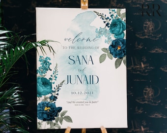 May include: A wedding announcement sign with a watercolor background and teal floral designs. The sign reads "Welcome to the wedding of Sana and Junaid 10.12.2021" and includes the quote "And We created you in pairs."