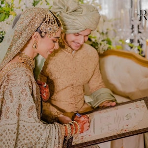 May include: A couple signing a document at a wedding ceremony. The bride is wearing a green and gold embroidered dress with a jeweled headpiece. The groom is wearing a beige sherwani and a green turban. The document is in a brown frame with a floral design.
