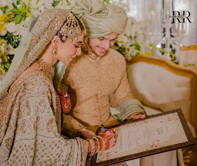 May include: A couple signing a document at a wedding ceremony. The bride is wearing a green and gold embroidered dress with a jeweled headpiece. The groom is wearing a beige sherwani and a green turban. The document is in a brown frame with a floral design.