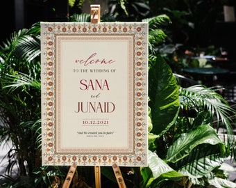May include: A wedding welcome sign on an easel. The sign has a cream background with a decorative border in brown and gold. The text reads "welcome to the wedding of SANA and JUNAID 10.12.2021". The sign is surrounded by lush green plants.