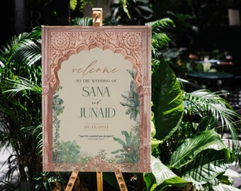 May include: A wedding welcome sign with a pink and green floral design. The sign reads "Welcome to the wedding of Sana and Junaid 10.12.2021" and includes a quote in Arabic script.