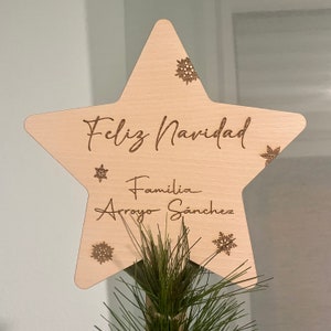Op de afbeelding: Een houten stervormige kerstboomtopper met de tekst "Feliz Navidad" en "Familia Arroyo Sanchez" in een cursief schrift. De ster is versierd met sneeuwvlokken.