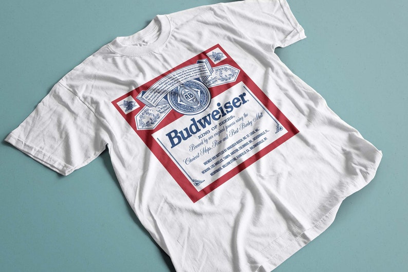 May include: White t-shirt with a red and blue Budweiser logo printed on the front. The logo features the text "Budweiser" in blue and "King of Beers" in smaller text below. The logo also includes a red and white crest with the letters "AB" in blue.