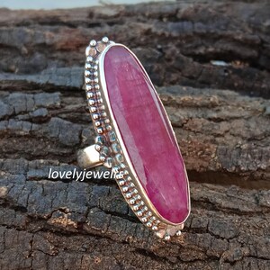 Puede incluir: Un anillo de plata con una gran gema ovalada de color rojo intenso. La gema está engastada en una banda de plata con un borde detallado de cuentas. El anillo se muestra sobre una superficie texturizada de color marrón oscuro. El texto "lovelyjewels" es visible.