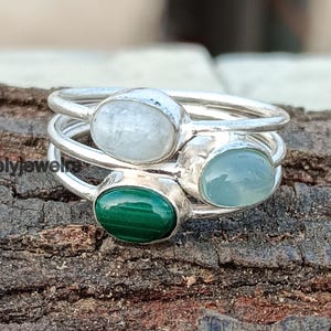 May include: A silver ring with three oval-shaped gemstones. The stones are a white moonstone, a light blue chalcedony, and a green malachite.