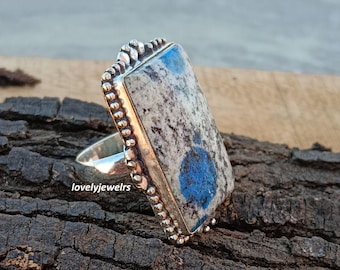 May include: A rectangular silver ring featuring a large, light-colored stone with blue and gray patterns. The ring has a detailed, beaded border and a wide band. The stone's surface is polished, and the ring is set against a dark, textured background.