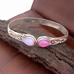 Op de afbeelding: Een zilverkleurige armband met twee druppelvormige roze en lavendelkleurige edelstenen. De armband heeft sierlijke details bij de edelstenen en de tekst "lovelyjewelrs". Het wordt gepresenteerd op een donkere houten ondergrond.
