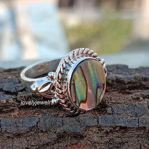 Puede incluir: Un anillo de plata con una incrustación redonda de concha de abulón. El anillo tiene un diseño detallado con una banda trenzada y un acento decorativo en el lateral.