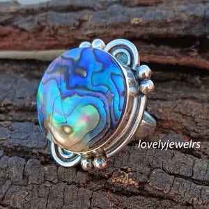 Pode incluir: Anel de prata com um grande cabochão de concha de abalone iridescente. A concha apresenta azuis, verdes e roxos vibrantes. O anel tem uma cravação de prata decorativa com detalhes ornamentados e o texto "lovelyjewelrs".