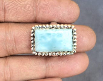 Könnte beinhalten: Ein rechteckiger Ring mit einem hellblauen Edelstein und einem silbernen Perlenrand. Der Edelstein hat ein wolkiges Aussehen. Der Ring wird in einer Hand vor grauem Hintergrund gehalten. Schmuck.