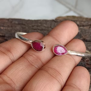 Può includere: Un bracciale a polsino argentato con due gemme rosse a forma di goccia. Il bracciale ha un design aperto, con le gemme incastonate a ciascuna estremità. Le gemme sono di un colore rosso vibrante e ricco, che aggiunge un tocco di colore alla fascia argentata.