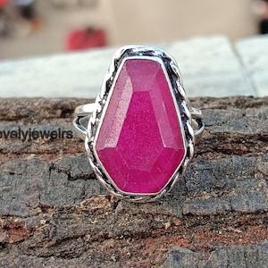 May include: A silver ring with a large, faceted, magenta-colored gemstone. The gemstone has a unique, elongated shape and is set in a detailed silver band. The ring is displayed on a textured wooden surface.