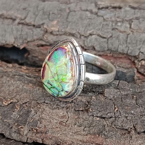 May include: A silver ring featuring a pear-shaped, iridescent gemstone with green, purple, and yellow hues. The stone has a cracked, textured appearance, set in a detailed silver bezel. The ring band is a simple, polished silver.