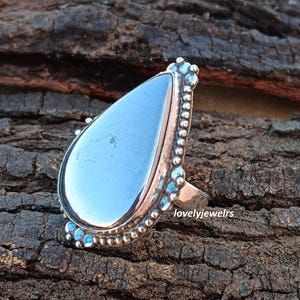 May include: A silver ring with a teardrop-shaped blue gemstone set in a beaded silver band. The ring is on a wooden surface.