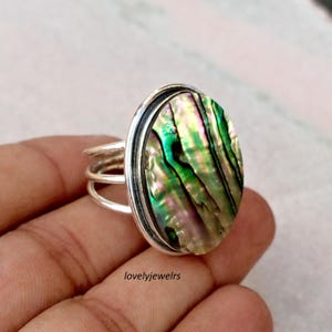 May include: Oval-shaped abalone shell ring with a silver band. The iridescent shell features green, pink, and purple hues. The ring has a triple-band design on the sides. The ring is a piece of jewelry.