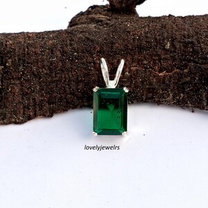 Puede incluir: Un colgante rectangular con talla esmeralda con una piedra preciosa verde vibrante engastada en una montura plateada. El colgante tiene una fianza simple para una cadena. El texto "lovelyjewelrs" es visible en la parte inferior de la imagen.