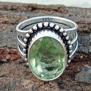 Pode incluir: Um anel de prata com uma grande pedra preciosa oval verde claro. O anel tem uma banda texturizada e uma moldura decorativa ao redor da pedra. O texto "925" é visível na banda. O anel está sobre uma superfície de madeira.