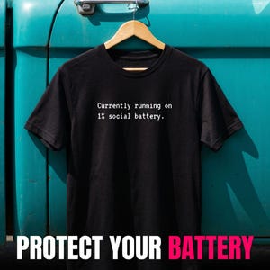 Könnte beinhalten: Ein schwarzes kurzärmeliges T-Shirt mit dem weißen Aufdruck "Currently running on 1% social battery." Das Shirt hängt an einem Holzkleiderbügel vor einem türkisfarbenen Hintergrund, mit dem Text "PROTECT YOUR BATTERY" darunter.