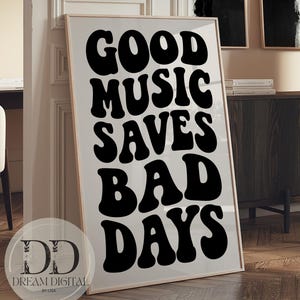May include: A rectangular framed wall art print featuring the black text GOOD MUSIC SAVES BAD DAYS in a bold, wavy, retro-inspired font against a light beige background. The print is encased in a thin, light-colored wooden frame.