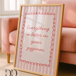 May include: A framed print with the text "Everything is better in your pyjamas" in red script. The print has a pink and white striped background and a red heart border. The frame is light brown.