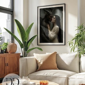 May include: A framed wall art print featuring a dark, moody illustration of a horned, dark-skinned figure embracing a person with long dark hair in a white dress. The artwork is displayed in a thin black frame, showcasing a romantic, gothic scene.