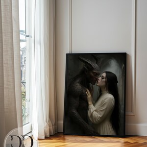 May include: A rectangular framed wall art print featuring a dark-skinned horned creature with glowing red eyes and a person with long dark hair wearing a cream-colored long-sleeved dress. The two figures are positioned face-to-face in a close embrace.