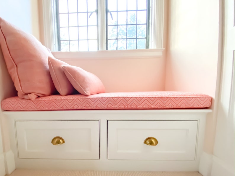 May include: A white window seat with two drawers and a pink cushion. The window cushion has a chevron pattern and is topped with two pink pillows.