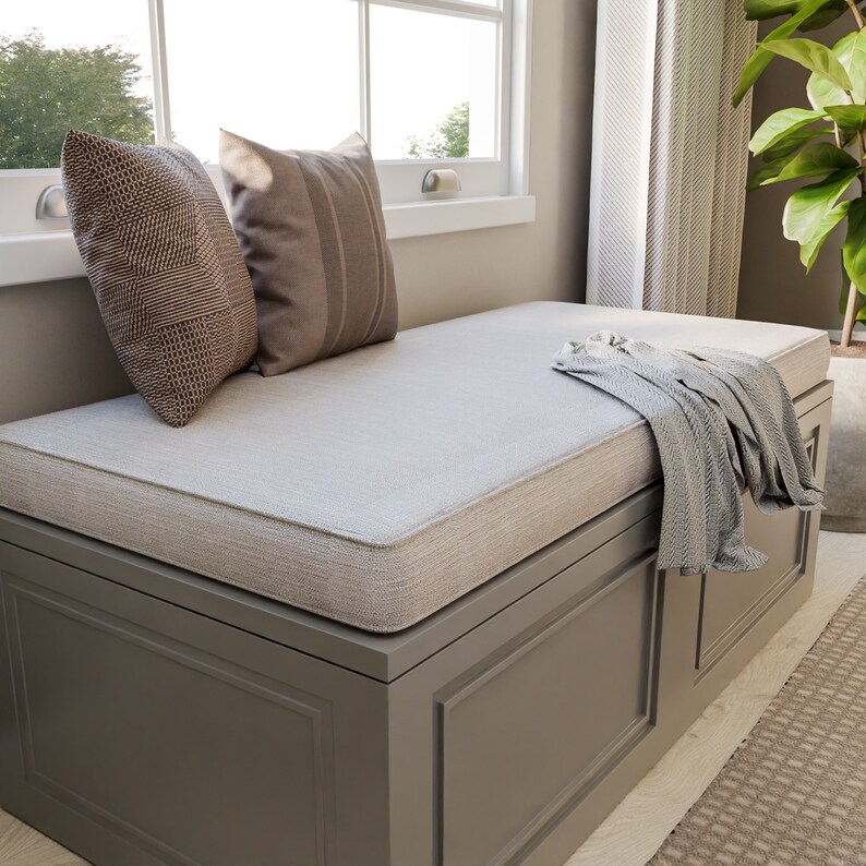 Puede incluir: Un asiento de ventana gris con un coj&iacute;n beige y dos almohadas marrones. Una manta gris est&aacute; colocada sobre el asiento. El asiento tiene un compartimento de almacenamiento con un marco de madera.