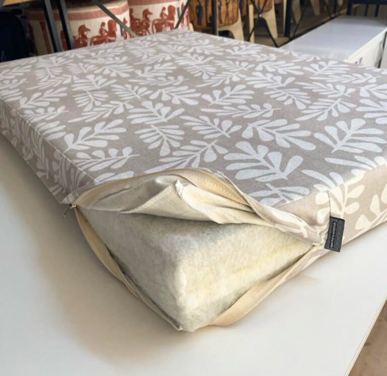 A beige cushion with a white leaf boho pattern. The cushion is zippered and has a removable, white foam insert.