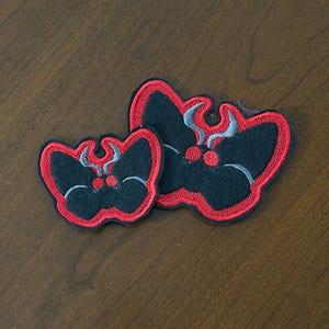May include: Two embroidered moth patches in different sizes, featuring a black body and wings outlined in vibrant red thread. Each moth has two prominent red circular eyes and curved grey antennae, creating a distinct insect shape.