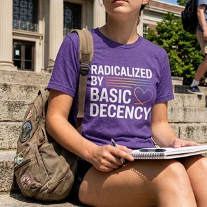 Op de afbeelding: Een paars t-shirt met de tekst "RADICALIZED BY BASIC DECENCY" in het wit, met een regenbooglijn en een hartje. De persoon draagt een bruine rugzak met patches en houdt een notitieboek en pen vast.
