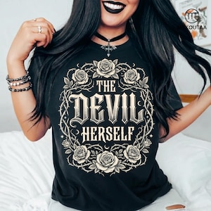 Puede incluir: Camiseta negra con la frase "THE DEVIL HERSELF" en fuente gótica, rodeada por una corona floral de rosas y vides. El diseño crema contrasta con la tela oscura. Una camiseta gráfica audaz y elegante.