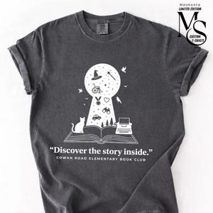 Peut inclure: T-shirt gris foncé avec un motif blanc en forme de trou de serrure rempli d'illustrations fantaisistes, dont un chapeau de sorcière, un chat et une machine à écrire. Le texte dit "Discover the story inside." et "COWAN ROAD ELEMENTARY BOOK CLUB."