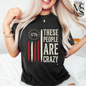 Peut inclure: T-shirt noir avec un motif de drapeau américain vieilli. Le t-shirt présente l'année "1776" et le texte "These People Are Crazy" en blanc. Conception de t-shirt personnalisée.