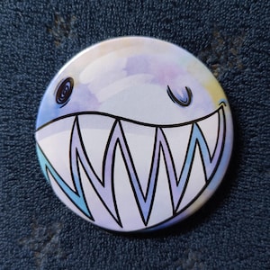 Pode incluir: Um broche redondo com uma ilustração em estilo aguarela de uma criatura com um sorriso largo e dentado. O design utiliza tons suaves de azul, roxo e amarelo, com contornos pretos fortes que definem os dentes afiados e os olhos.