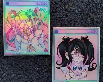 Puede incluir: Dos pegatinas holográficas con ilustraciones de estilo anime. Una muestra a una chica con coletas coloridas y una expresión angustiada. La otra representa a una chica con coletas negras sosteniendo una criatura alada triste. Ambas tienen un marco similar a una ventana.