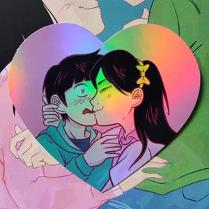Può includere: Un adesivo olografico a forma di cuore con un'illustrazione stilizzata di due persone che si abbracciano. Una persona ha i capelli scuri con un fiocco giallo, l'altra ha i capelli scuri e una felpa con cappuccio color ottanio. Finitura iridescente.