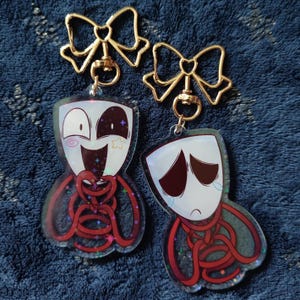 Puede incluir: Dos llaveros con personajes de dibujos animados. Uno muestra un personaje feliz con una estrella y una espiral, el otro un personaje triste con lágrimas. Ambos tienen máscaras blancas y cuerpos rojos en forma de cuerda. Llaveros dorados en forma de lazo.
