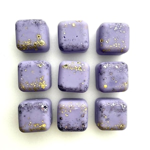 Puede incluir: Nueve ceras aromáticas cuadradas de color lavanda con bordes redondeados dispuestas en una cuadrícula. Cada cera tiene motas doradas y plateadas. El fondo es blanco.