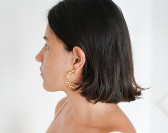 May include: A woman with short, dark hair wearing a white, sleeveless top and gold hoop earrings.