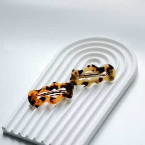 Op de afbeelding: Twee haarclips met een golvend schildpadpatroon, in bruin- en beigetinten, liggen op een wit, gewelfd, gestructureerd oppervlak. De clips hebben een goudkleurige metalen sluiting. Haaraccessoires.