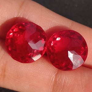 Puede incluir: Dos piedras preciosas redondas y rojas con un corte facetado. Las piedras preciosas están en un dedo.
