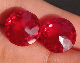May include: Two round, red gemstones with a faceted cut. The gemstones are on a finger.
